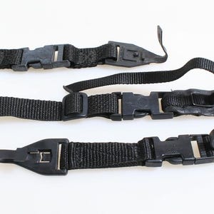 May include: Three black camera straps with plastic buckles and adjustable nylon webbing. The straps are laid out on a white surface, showcasing their design and functionality for securing a camera.