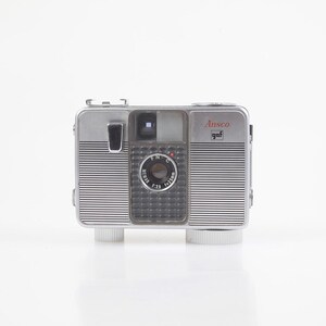 May include: A vintage silver Ansco camera with a black lens and a red "Ansco" logo. The camera has a rectangular shape with a ribbed design and a clear lens cover. The camera is mounted on two white cylindrical feet.
