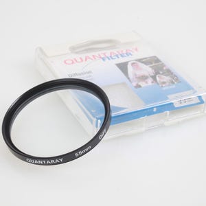May include: A black Quantaray 55mm Diff filter for a camera lens. The filter is circular with white text and sits next to its clear plastic packaging. The packaging has a photo of a bride and groom.