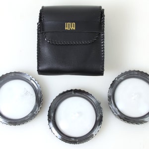 May include: Three black camera lens filters with a white interior and a black leather case with the word "HOYA" printed in gold letters.