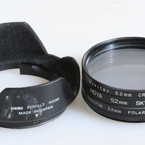 Może przedstawiać: Czarna osłona przeciwsłoneczna Sigma Perfect Hood i zestaw filtrów do obiektywu aparatu 52 mm. Zestaw filtrów zawiera filtry Vivitar, Hoya i Tokina, w tym filtr krzyżowy, skylight i polaryzacyjny. Osłona przeciwsłoneczna jest oznaczona "Made in Japan".
