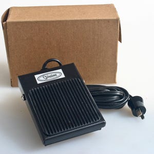 May include: Black Spiratone foot switch with a ribbed surface, a handle, and a power cord with a plug. The switch is rectangular and sits in front of a brown cardboard box.