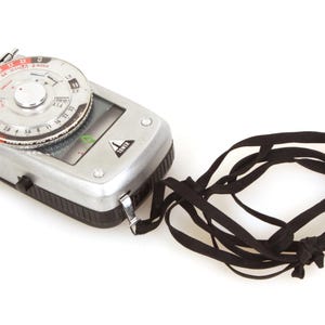 May include: A vintage silver light meter with a black and silver dial and a black strap. The light meter has a black and silver body with a dial on top. The strap is made of black fabric.