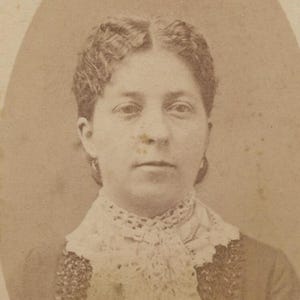 Op de afbeelding: Een sepia-kleurige vintage portretfoto van een persoon met golvend haar in een middenscheiding. Het onderwerp draagt een kledingstuk met een hoge kraag en een gedetailleerd, wit kanten halsstuk of jabot met een gestructureerd patroon.