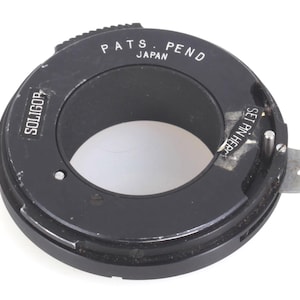 Puede incluir: Un adaptador de lente Soligor negro con el texto "PATS. PEND JAPAN" y "SOLIGOR" impreso en el anillo exterior. El adaptador tiene una abertura circular y una palanca de metal plateado. También se ven las palabras "SET PIN HERE".