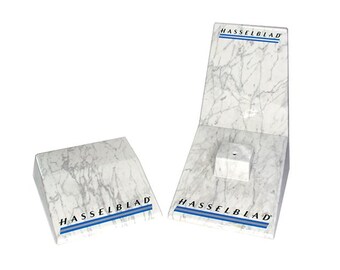 Könnte beinhalten: Zwei Displayständer mit Marmormuster und dem blauen Text "HASSELBLAD". Einer der Ständer ist abgewinkelt und hat einen kleinen Würfel auf der Basis. Der andere ist ein flaches, rechteckiges Stück. Beide haben ein weißes und graues Adernmuster.