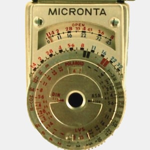 May include: A vintage Micronta light meter with a silver metal casing. The device features multiple dials with numbers and markings for settings like ASA, DIN, and LVS. The word "MICRONTA" is printed at the top.