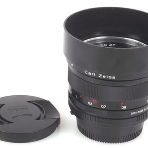 Może przedstawiać: Czarny obiektyw do aparatu Carl Zeiss z osłoną obiektywu. Obiektyw ma czarną obudowę z białym napisem "Carl Zeiss" i "Lens made in Japan". Osłona obiektywu jest czarna z logo Zeiss.