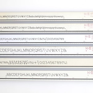May include: A set of vintage Leroy lettering guides, featuring various fonts and character sets. The guides are cream-coloured with black lettering and a ruler along the bottom. The K&E Keuffel & Esser Co. logo is visible on each guide.