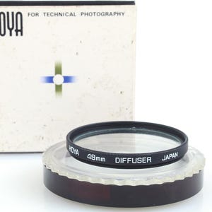 May include: A Hoya 49mm diffuser filter with a black ring and the text "DIFFUSER JAPAN" is displayed on a clear plastic base with a dark brown rim. The box behind it reads "HOYA FOR TECHNICAL PHOTOGRAPHY".