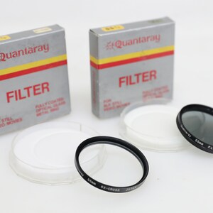 May include: Two unopened boxes of Quantaray 55mm filters for SLR still and video movies. The boxes are grey with red and yellow accents. The text on the boxes reads "Quantaray" and "Filter". There are two clear plastic cases with white inserts and a black 55mm filter.