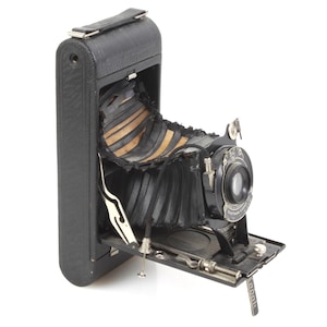 Op de afbeelding: Een vintage zwarte vouwcamera met een balg en een lens. De camera is open en de balg is uitgetrokken.