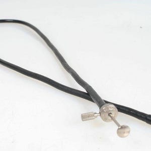May include: A black, flexible cable with a metal fitting and a small, round, silver-colored knob. The cable is lying on a white surface. The cable appears to be a camera accessory.