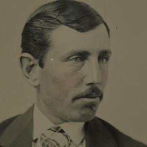 Puede incluir: Un retrato en tonos sepia de un hombre con bigote, que lleva una chaqueta oscura y una corbata estampada. El hombre tiene el pelo corto y bien peinado y una expresión seria. La imagen es una fotografía antigua.