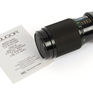 Op de afbeelding: Een zwarte Soligor multi-coated zoom-macro C/D auto lens met instructies in meerdere talen. De lens heeft een variabele brandpuntsafstand en macro-mogelijkheden.