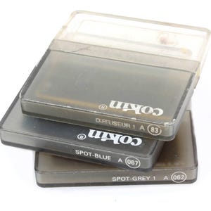 May include: Three Cokin brand camera filters in their original cases. The filters are labeled "DIFFUSEUR 1 A (83)", "SPOT-BLUE A (067)", and "SPOT-GREY 1 A (062)". The cases are gray and translucent, stacked on top of each other.