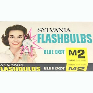 May include: A box of Sylvania Blue Dot M2 flashbulbs. The box features a woman holding a camera and a flashbulb. The text on the box reads "Sylvania Flashbulbs Blue Dot M2 Twelve Clear Bulbs".