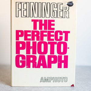 Könnte beinhalten: Ein weißes Buch mit dem Titel "FEININGER THE PERFECT PHOTOGRAPH" in schwarzer und pinkfarbener Schrift. Das Buch ist im Verlag AMPHOTO erschienen.