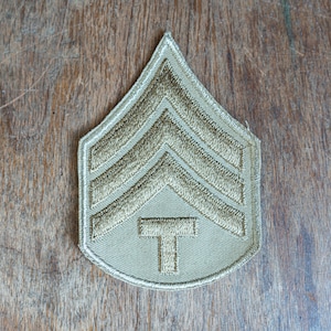 May include: A beige military rank patch with three gold embroidered chevrons and a gold embroidered 'T' in the center.