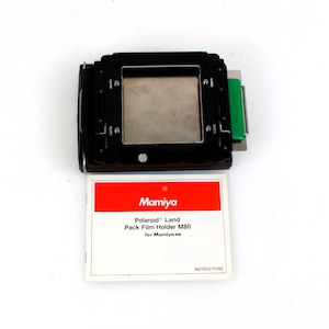 May include: A black Mamiya Polaroid Land Pack Film Holder M80 for Mamiya cameras. The holder has a silver interior and a green component on the side. A white card with red accents and the Mamiya logo is below the holder.
