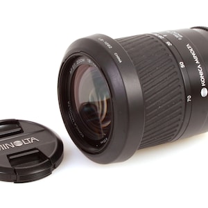 May include: A black Konica Minolta AF DT Zoom 18-135mm lens with a black lens cap. The lens has a silver band with the text "Konica Minolta AF DT Zoom 18-135mm" and "Made in China".