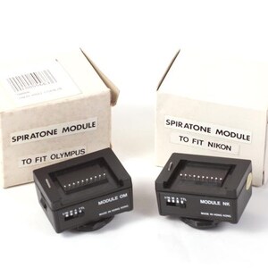 May include: Two black Spiratone camera modules, one labeled "TO FIT OLYMPUS" and the other "TO FIT NIKON", sit in front of their respective white boxes. The boxes are labeled "SPIRATONE MODULE".