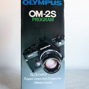Puede incluir: Una cámara Olympus OM-2S Program negra y plateada con un objetivo plateado. La cámara está en un folleto en blanco y negro con el texto "The OLYMPUS Program Camera that's Designed for Unlimited Growth."