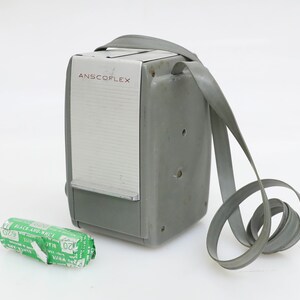 May include: A gray Anscoflex camera with a black and white film roll. The camera has a strap and a white front panel with the word "Anscoflex" printed in red.