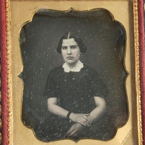 Op de afbeelding: Een vintage zwart-wit portret van een persoon in een donkere jurk met een witte kanten kraag. Het portret is omlijst in een decoratieve gouden en rode lijst, met een historische foto.