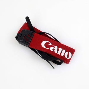 Könnte beinhalten: Roter Canon Kameragurt mit weißem Schriftzug. Der Gurt hat eine schwarze Kunststoffschnalle und schwarze Nylongurte. Das Canon-Logo ist auf dem roten Gurt zu sehen.
