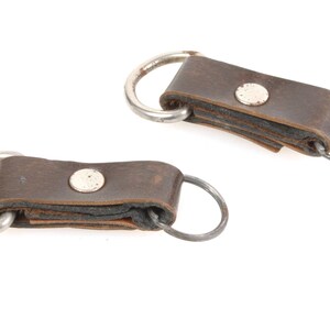 May include: Two brown leather keychains with silver-colored metal rings and rivets. The leather is worn, showing signs of use. The keychains are set against a white background.