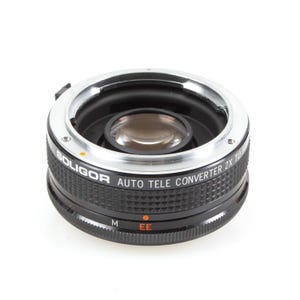May include: A black and silver Boligor Auto Tele Converter 2X TO 100mm lens adapter. The adapter has a ridged black body with silver accents and a lens element visible in the center. The text "BOLIGOR AUTO TELE CONVERTER 2X TO 100mm" is printed on the side.