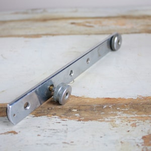 Puede incluir: Una barra de metal plateada con dos botones ajustables y cinco agujeros. La barra está diseñada para ser utilizada como un soporte de montaje para una cámara u otro equipo.