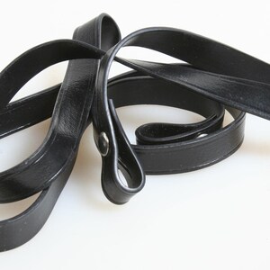 May include: A black leather strap with a silver-tone rivet. The strap is looped and curved, lying on a white surface. The strap appears to be for a bag or purse.