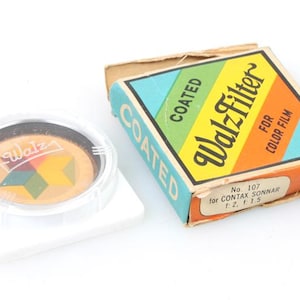May include: A vintage camera lens filter in a white plastic case. The filter is labeled "Walz Filter" and is coated. The box is labeled "No. 107 for Contax Sonnar f:2 1:15 Type F (85C)".