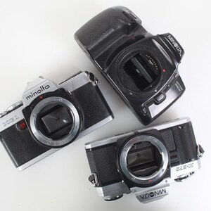May include: Three vintage Minolta SLR cameras are arranged on a white surface. The cameras are black and silver, with visible lenses and the Minolta brand name. The X-370 and X-700 models are visible.