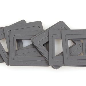 Peut inclure: Plusieurs montures de diapositives grises, chacune avec une ouverture rectangulaire. Les montures sont empilées et légèrement superposées, créant un effet de superposition. Les articles sont en matériau mat, sur fond blanc.