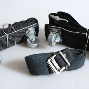 May include: Black nylon webbing straps with metal casters and buckles. Two sets of straps are bundled with rubber bands, and one strap is rolled up with a metal buckle. The casters are silver.
