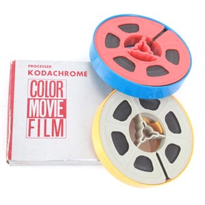 Pode incluir: Natureza morta de bobinas de filme vintage e uma caixa de Kodachrome Color Movie Film. As bobinas são azuis, vermelhas e amarelas com filme preto no interior. A caixa é prateada com texto vermelho e branco.