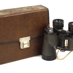 Könnte beinhalten: Schwarzes Fernglas mit brauner Ledertasche. Das Fernglas hat schwarze Okulare und ein silbernes Fokusrad. Die rechteckige Tasche hat einen Griff und einen silbernen Verschluss.
