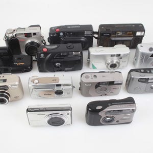 May include: A collection of various point-and-shoot film cameras in silver, black, and gray. The cameras are arranged on a white surface, showcasing different designs and brands, including Kodak and Vivitar.