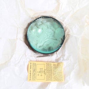 Könnte beinhalten: Ein Vintage-Kondensatorsatz mit einer kreisförmigen, teal-farbenen Linse und einem schwarzen Rand auf zerknittertem, weißem Papier. Ein kleines gelbes Anleitungsblatt ist darunter zu sehen, mit Montageanweisungen.