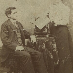 May include: Sepia-toned portrait of a man seated and a woman standing. The man wears a dark suit, waistcoat, and tie, seated on a chair. The woman wears a white blouse and dark skirt. A small table with a patterned cloth is between them.