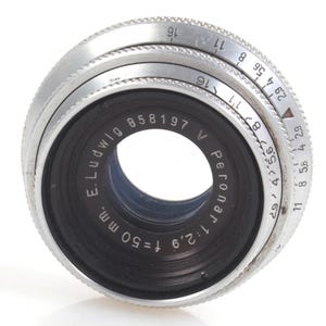 May include: Close-up of a vintage camera lens with a black center and silver rings. The lens has the text "E. Ludwig Peronar 1:2.9 f=50 mm." and a serial number. The aperture settings are visible on the silver ring.
