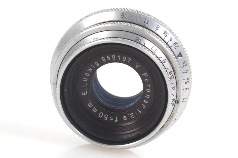May include: Close-up of a vintage camera lens with a black center and silver rings. The lens has the text "E. Ludwig Peronar 1:2.9 f=50 mm." and a serial number. The aperture settings are visible on the silver ring.