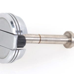 Puede incluir: Un tapón de botella plateado con una cabeza circular y un vástago cilíndrico. La cabeza tiene un diseño segmentado con tres ranuras paralelas. Se ve una pieza de goma negra.