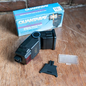 May include: A black Quantaray QRF-500DX wireless remote flash with a blue ring around the flash head. The flash is on a wooden surface with its box and accessories.