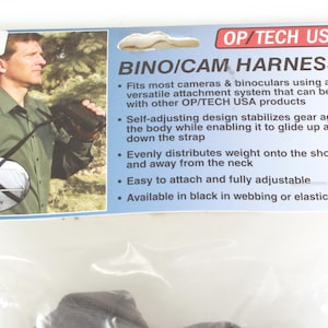 May include: OP/TECH USA Bino/Cam Harness package. The package features a man wearing the harness with binoculars. The text on the package says the harness fits most cameras and binoculars and is available in black in webbing or elastic.