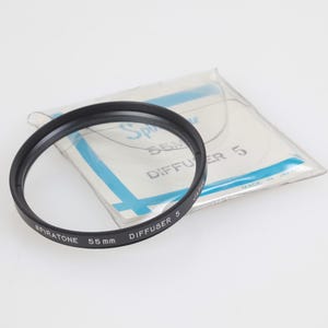 May include: A black Spiratone 55mm Diffuser 5 camera lens filter. The filter is circular with white text. A clear plastic package with blue accents and text is behind the filter.