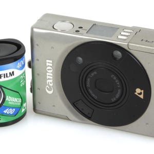 May include: A silver Canon ELPH digital camera with a black lens and a roll of Fujifilm 400 film. The film canister is black and green, with text indicating 24mm film. The camera has a zoom lens and a small display.